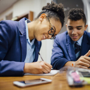 A school girl sitting at a desk writing on paper with a school boy sitting next to her