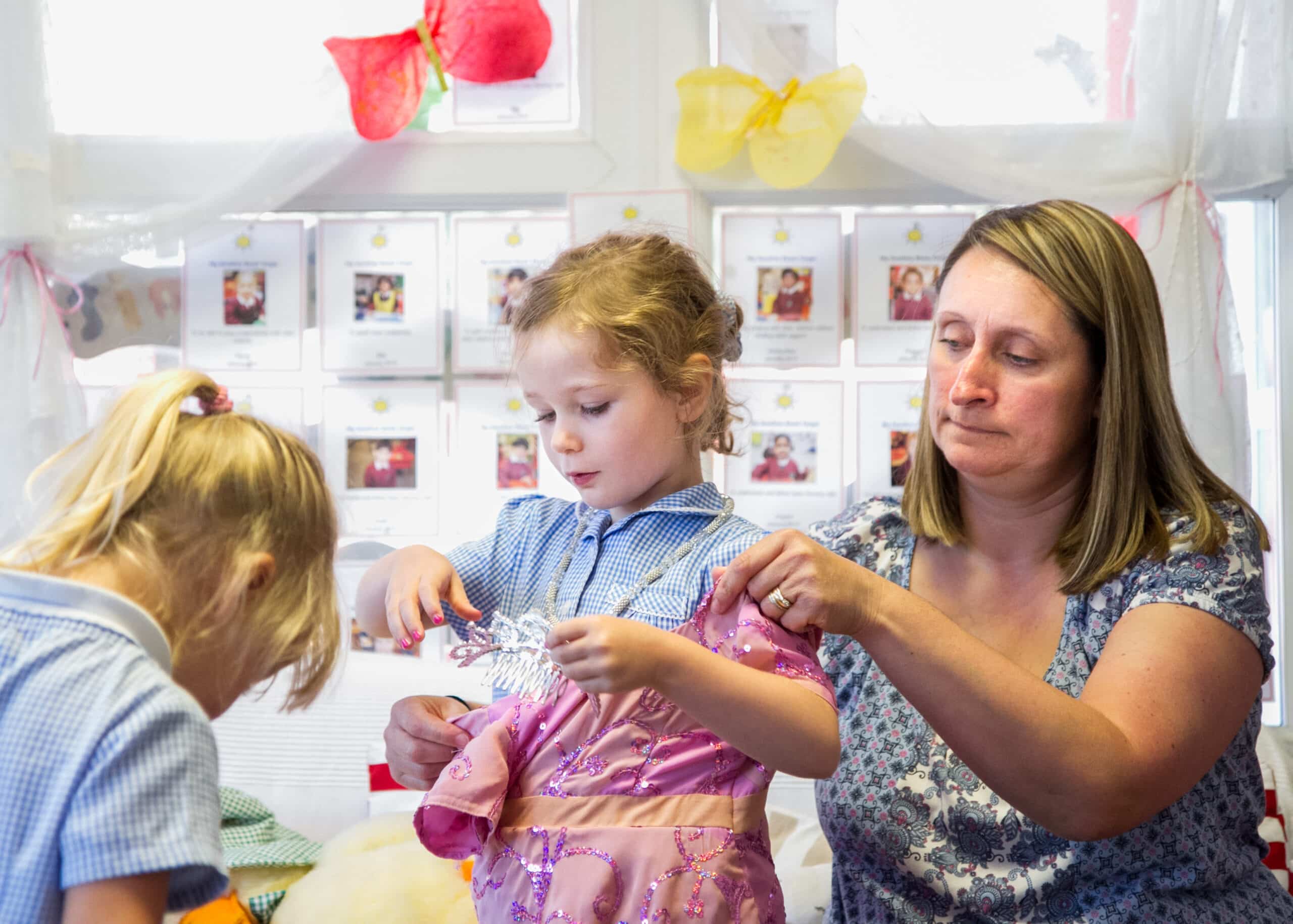 Two young children are shown at school, perhaps in a nurture group, crafting. One girl is cutting something out using scissors and the teacher is next to her, watching over.