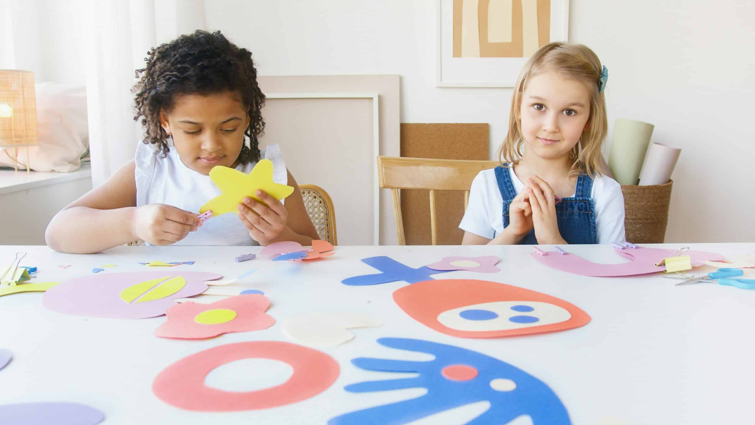 Two young children are sat at a table together. Colourful pieces of paper are shown in front of them in a variety of shapes. One girl is cutting up a yellow piece of paper, the other looks at the camera and smiles.