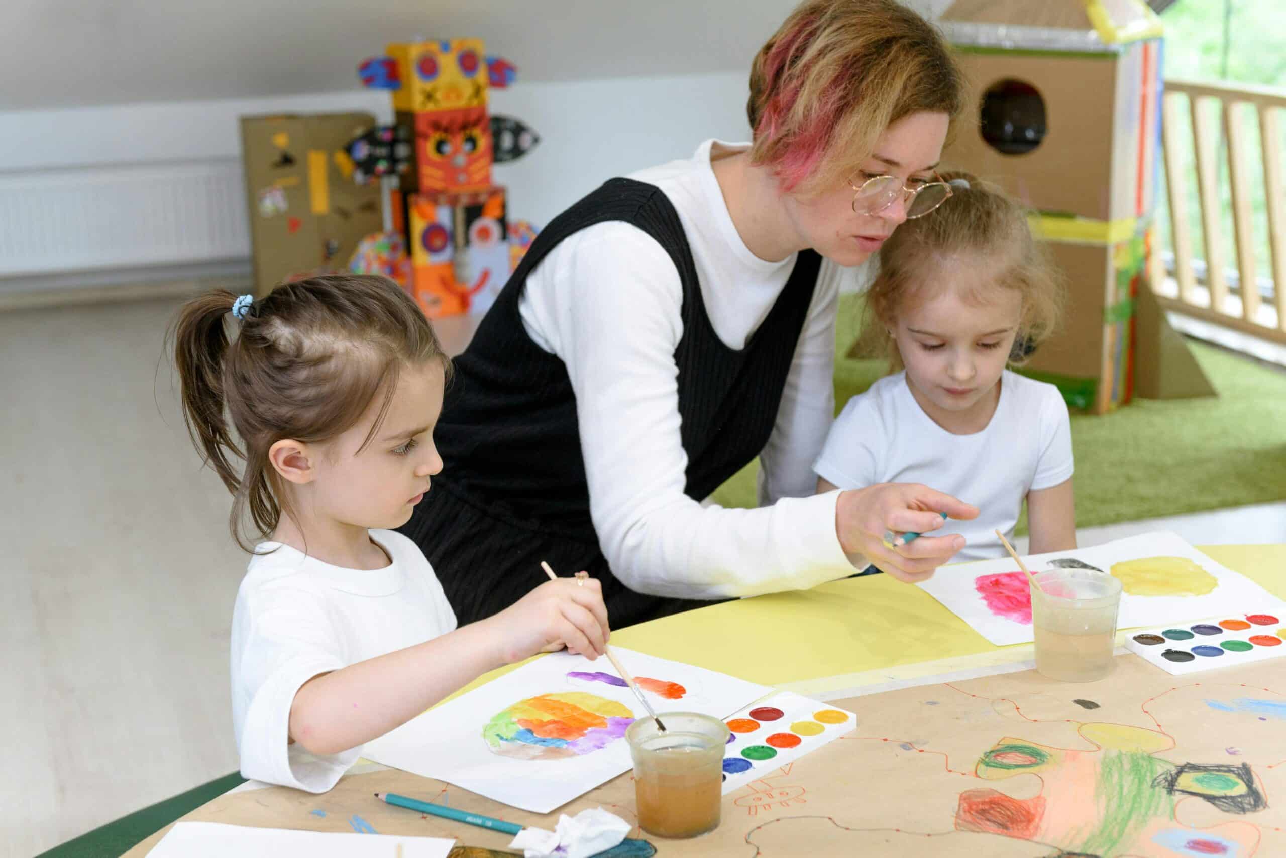 Two children are shown at a table with a teacher inbetween them. They are both using paint and paintbrushes on paper.