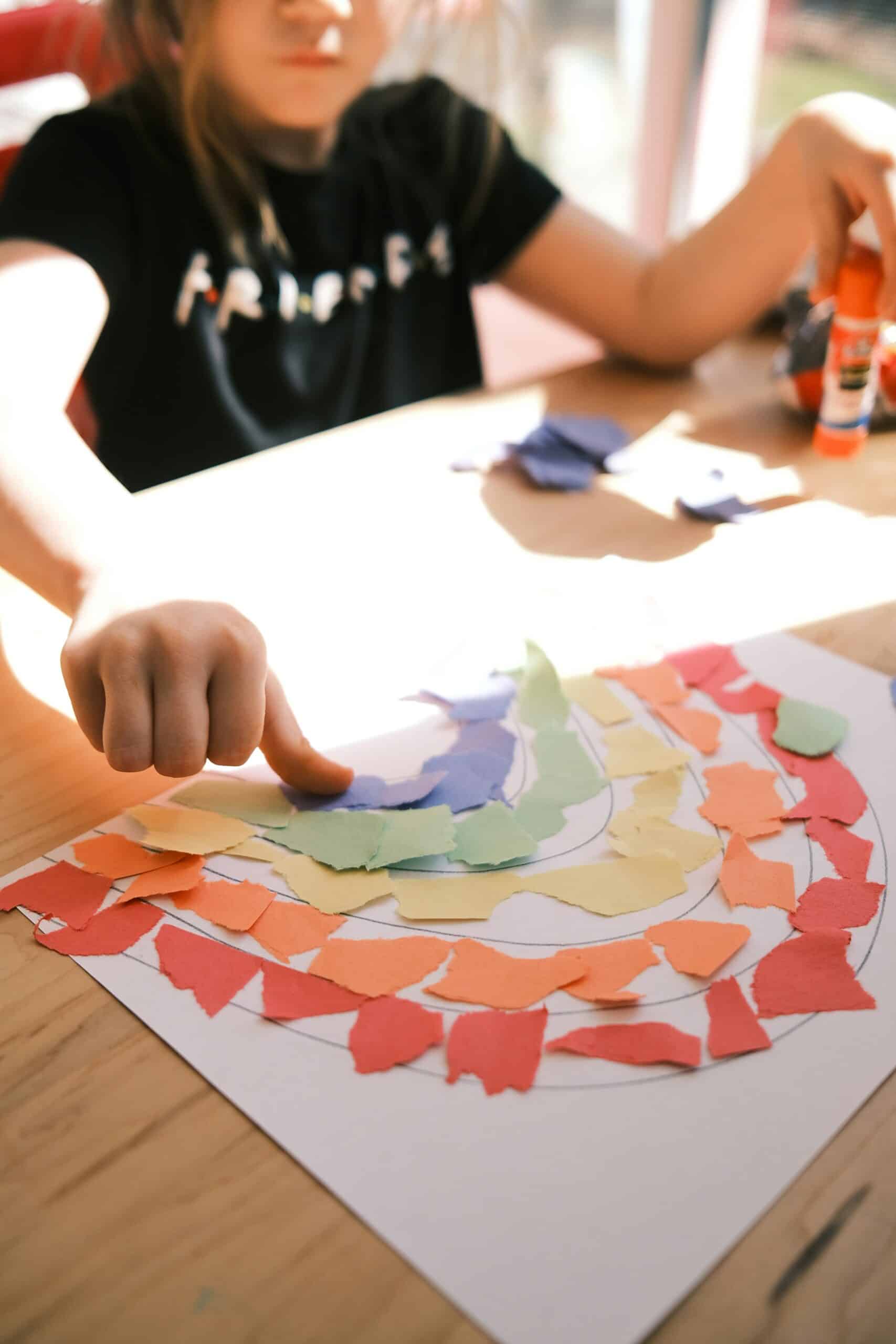 A child is shown from the shoulders down crafting something at a table. There is paper in front of her with lots of colourful bits of paper cut out in the shape of a rainbow.