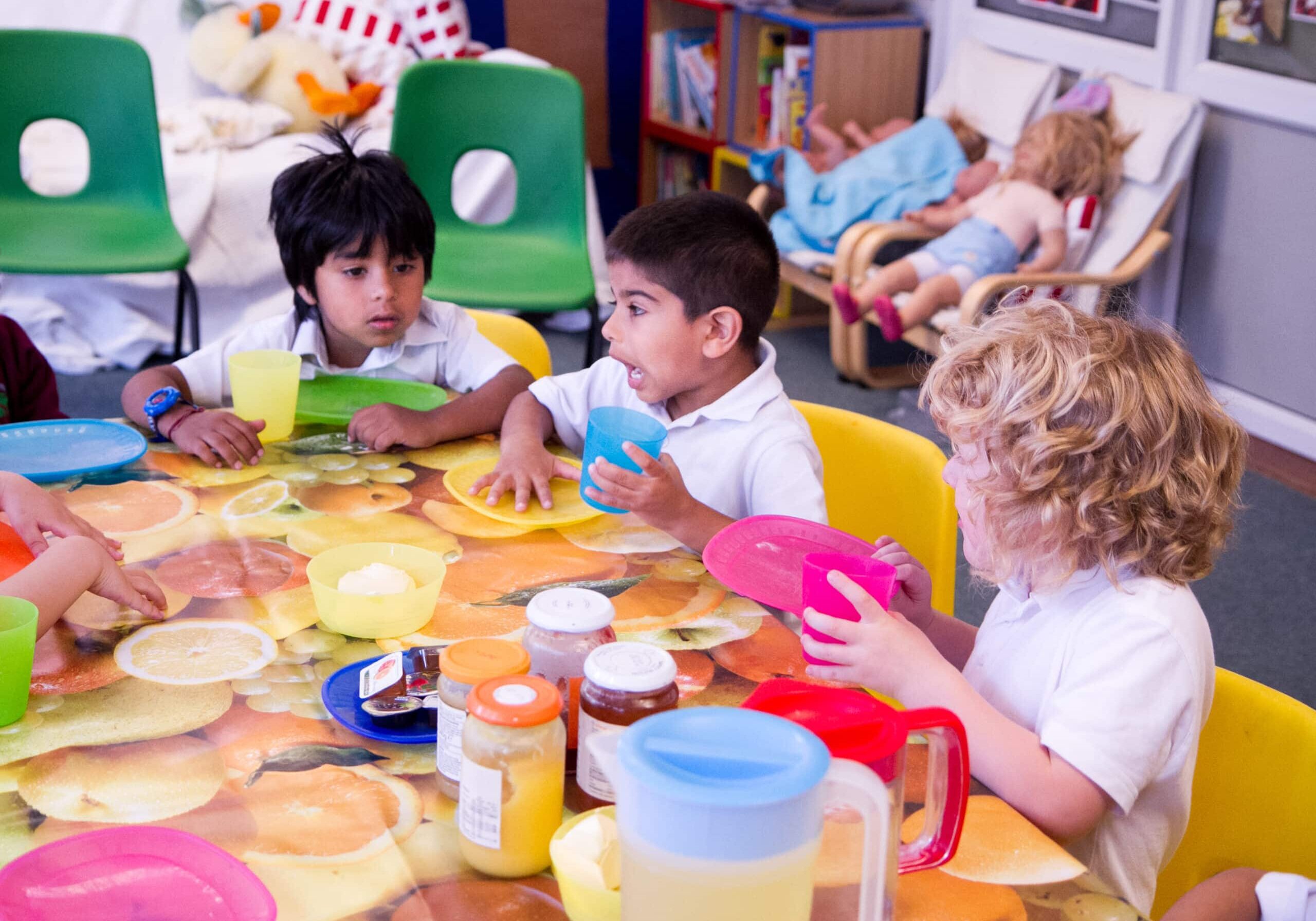 Three children sit together at a table in a nurture group. They have lots of plates and objects in front of them, perhaps ready to craft something. They look happy and engaged.